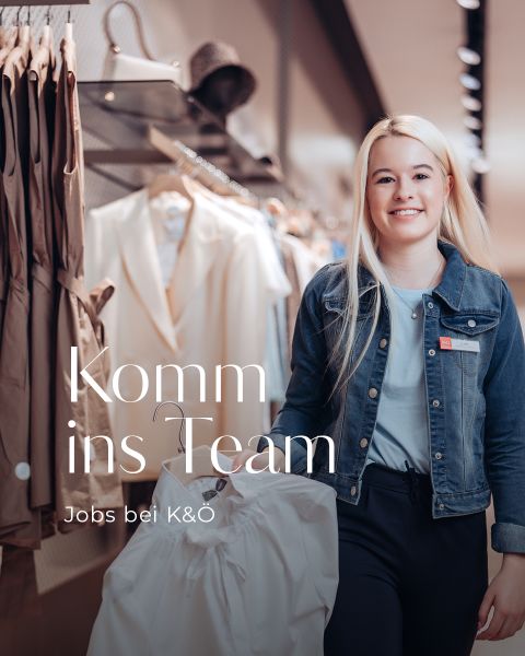 Eine junge Frau mit langen blonden Haaren, einer Jeansjacke und einer dunkelblauen Hose, hält in einem Bekleidungsgeschäft ein weißes Hemd in der Hand. Der Text lautet: „Komm ins Team, Jobs bei K&Ö.“ Im Hintergrund sind verschiedene Kleidungsstücke und Taschen zu sehen.