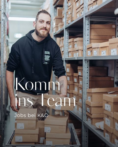 Eine Person mit Bart und schwarzem Kapuzenpullover steht in einem Gang, umgeben von Regalen voller Kartons. Der Text lautet: „Komm ins Team Jobs bei K&Ö.“