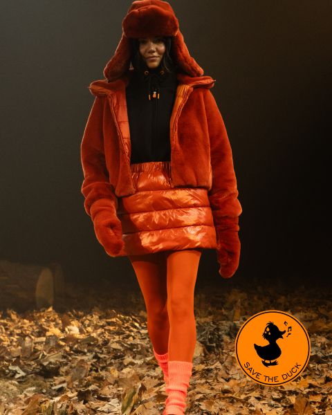 Ein Model läuft auf einem mit Blättern bedeckten Pfad. Es trägt eine orangefarbene Pelzjacke, einen passenden Rock und eine Mütze mit orangefarbenen Strumpfhosen und pfirsichfarbenen Socken. In der Ecke ist das Logo von „Save the Duck“ zu sehen.