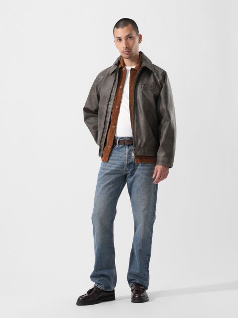 Vor einem schlichten Hintergrund steht ein Mann, der eine braune Jacke über einem Hemd, blaue Jeans und dunkle Schuhe trägt.