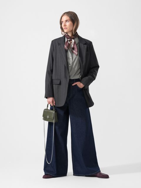 Eine Frau in einer Jeans mit weitem Bein, einem karierten Hemd, einem dunklen Blazer und einem Schal, die eine kleine grüne Handtasche in der Hand hält, steht vor einem schlichten weißen Hintergrund.