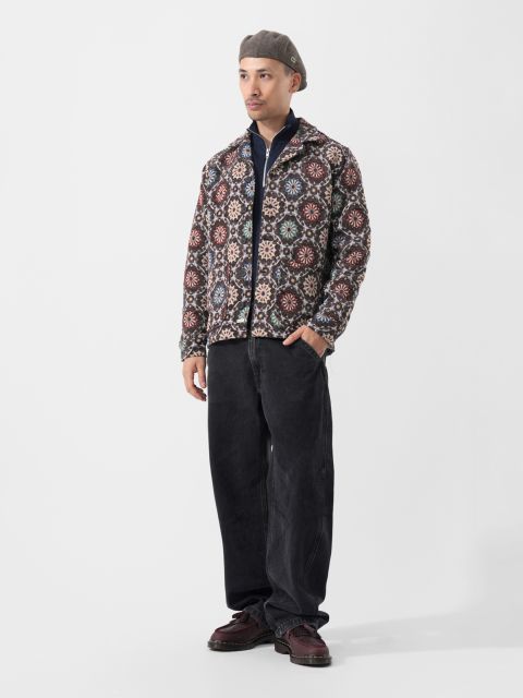 Ein Mann in einer gemusterten Jacke, einer schwarzen Hose, weinroten Schuhen und einer grauen Baskenmütze steht vor einem schlichten weißen Hintergrund.