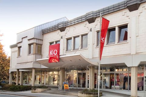 Das moderne Gebäude mit beiger Fassade beherbergt die Geschäfte K&Ö Kastner & Öhler und Giga X Sport. Rote und orangefarbene Schilder sind auffällig. Große Fenster und eine Straße davor unter klarem Himmel runden das Bild ab.