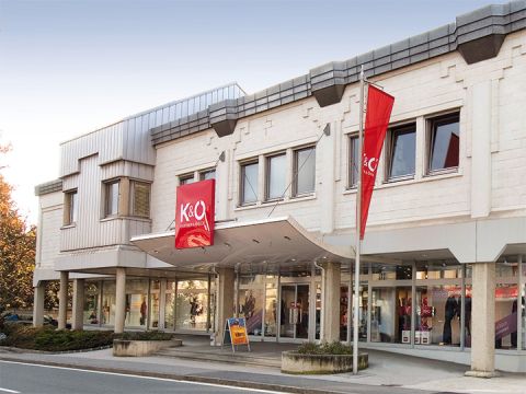 Das moderne Gebäude mit beiger Fassade beherbergt die Geschäfte K&Ö Kastner & Öhler und Giga X Sport. Rote und orangefarbene Schilder sind auffällig. Große Fenster und eine Straße davor unter klarem Himmel runden das Bild ab.