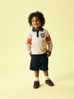 Ein Junge mit blondem Haar trägt eine schwarze Mütze, eine cremefarbene Fleecejacke über einem gestreiften Hemd, graue Jeans und Turnschuhe. Er steht vor einem einfarbig blauen Hintergrund und blickt mit gefalteten Händen nach unten.