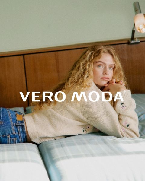 Eine Frau mit Brille und einem gerüschten Blumenkleid lehnt an einer weißen Wand vor einem blau gekachelten Hintergrund. Der Schriftzug "VERO MODA" erscheint in der Mitte des Bildes.