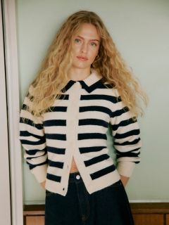 Eine Frau mit lockigem Haar steht an einer gekachelten Wand. Sie trägt eine hellgraue, wuschelige Strickjacke, schwarze, hoch sitzende Ledershorts und eine schwarze Strumpfhose, die Hände in den Taschen und einen selbstbewussten Gesichtsausdruck.