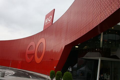 Ein modernes Gebäude mit einer großen, geschwungenen roten Fassade mit den Buchstaben „eo“. Auf dem Dach sind Schilder für K&O und Giga Sport zu sehen. Der Eingang ist aus Glas, daneben stehen Topfpflanzen, und im Vordergrund verläuft eine Kurve.