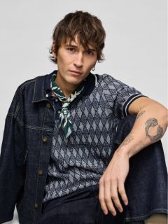 Ein junger Mann in Jeansjacke und Jeans kniet nieder und schaut zur Seite. Er trägt weiße Turnschuhe und hat eine sichtbare Tätowierung auf seinem Unterarm. Der Hintergrund ist einfarbig hellgrau.