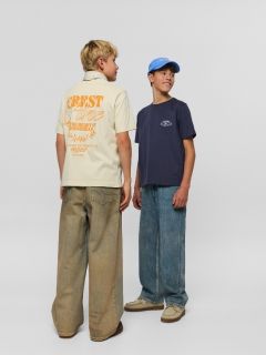 Zwei Jungen in Jeans und Jeanshemden posieren zusammen. Der eine sitzt auf einem Hocker, hat die Hände in den Schoß gelegt und einen Schal um den Hals, während der andere mit dem Arm auf dem Hocker steht und eine Mütze trägt.