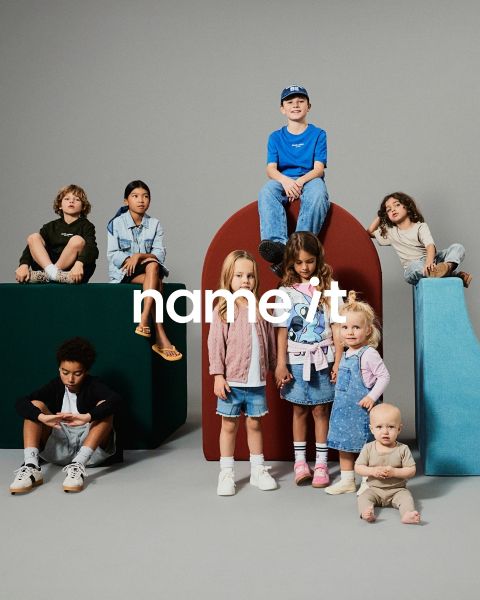 Eine Gruppe von acht verschiedenen Kindern steht vor einem grauen Hintergrund zusammen, alle tragen lässige, aufeinander abgestimmte Outfits in neutralen und Pastellfarben. Der Text "name it" erscheint in der Mitte des Bildes.