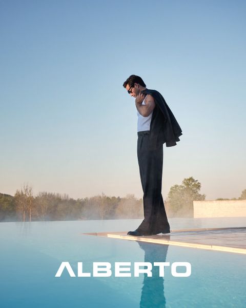 Ein Mann mit Brille, einem beigen Pullover mit Reißverschluss und einer hellen Hose sitzt auf einem Steinvorsprung am Meer. Er blickt zur Seite, unter ihm steht in weißer Schrift "ALBERTO". Der Hintergrund ist wolkenverhangen und ein Pier ist zu sehen.