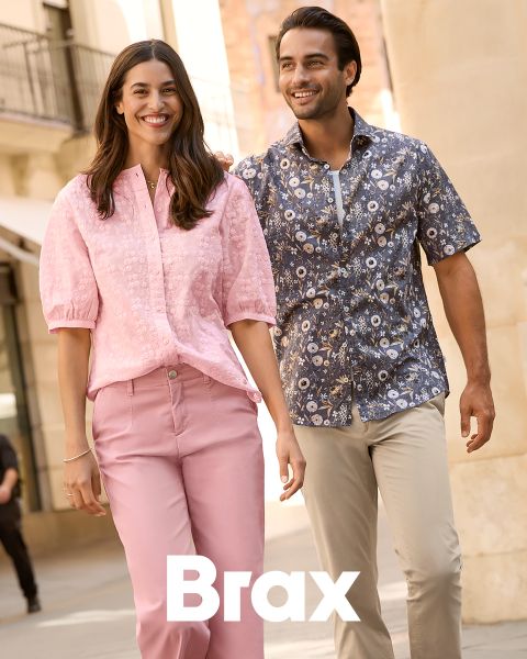 Eine Frau in einem gemusterten Mantel und einem roten Pullover steht neben einem Mann in einem marineblauen Wintermantel und einer beigen Hose. Sie lehnen an einer Steinmauer im Freien. Das Wort "Brax" erscheint am unteren Rand des Bildes.