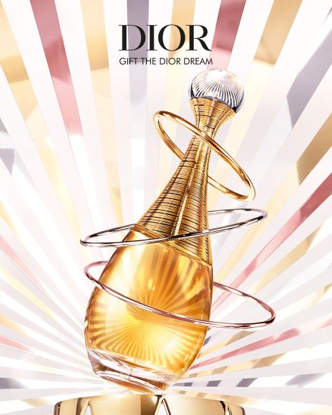 Ein goldener Parfümflakon mit eleganten Ringen und einer mit Juwelen besetzten Kappe steht vor einem leuchtenden Hintergrund mit Sonnenstrahlenmustern. Oben ist das Wort "DIOR" zu lesen.