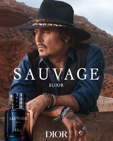 Ein Mann mit Hut und Schmuck lehnt sich an einen Holzzaun in einer Wüstenlandschaft mit roten Felsen. Im Vordergrund ist ein Flakon des Parfums Dior Sauvage Elixir zu sehen. Der Text "Sauvage Elixir Dior" ist überlagert.