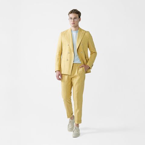 Ein Mann mit Sonnenbrille, einem hellrosa gemusterten Kurzarmhemd, weißen Hosen und grauen Turnschuhen steht vor einem schlichten weißen Hintergrund und hält eine weiße Jacke über der Schulter.