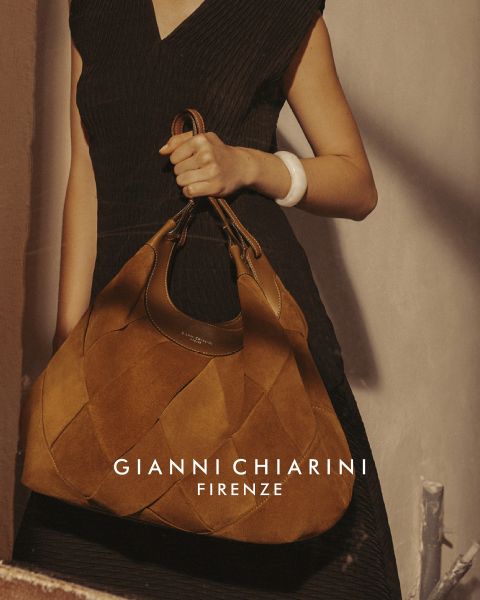 Person, die einen langen schwarzen Trenchcoat aus Leder und eine Sonnenbrille trägt und eine kleine Tasche mit Leopardenmuster hält. Der Hintergrund ist grau, mit dem Text „GIANNI CHIARINI FIRENZE“ in Weiß über dem Bild.