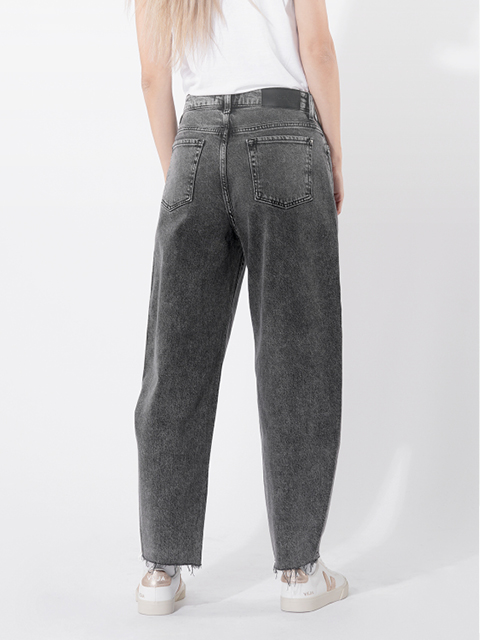 Eine Person, die eine hoch taillierte, locker sitzende, verwaschene schwarze Jeans mit den Händen in den Taschen trägt, gepaart mit einem weißen T-Shirt und weißen Turnschuhen, steht vor einem schlichten hellen Hintergrund.