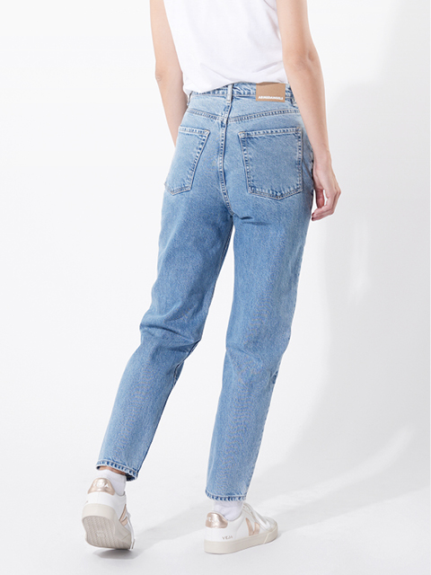 Eine Person, die hellblaue Jeans mit hoher Taille, ein weißes T-Shirt und weiße Turnschuhe trägt, steht vor einem schlichten weißen Hintergrund. Ihre linke Hand ruht auf ihrer Hüfte, während ihr rechter Arm an ihrer Seite herunterhängt.