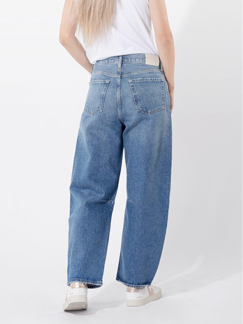 Eine Person, die eine locker sitzende blaue Jeans, ein weißes T-Shirt und weiße Turnschuhe trägt, steht mit einer Hand in der Tasche vor einem schlichten weißen Hintergrund.