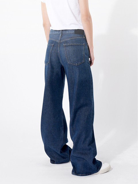 Eine Person, die eine weite Bluejeans und ein weißes T-Shirt trägt, steht vor einem einfarbig weißen Hintergrund. Nur die untere Hälfte des Körpers ist zu sehen; die Jeans ist weit geschnitten und verdeckt den größten Teil der Schuhe.