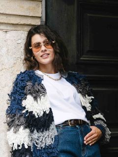 Eine Frau mit kurzen braunen Haaren, die eine Sonnenbrille, eine dicke blau-weiße Strickjacke, ein weißes T-Shirt und eine blaue Jeans trägt, steht lässig vor einer Steinmauer und einer dunklen Tür und lächelt selbstbewusst.