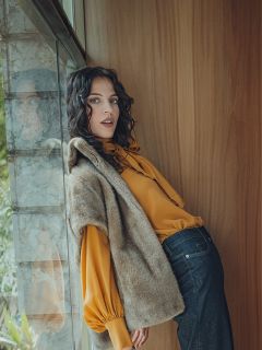Eine Frau mit gewelltem dunklem Haar lehnt an einer Holzwand an einem Fenster. Sie trägt eine gelbe Bluse mit Puffärmeln, eine braune Fellweste und dunkle Jeans. Ihr Gesichtsausdruck ist entspannt, während sie in die Kamera blickt.