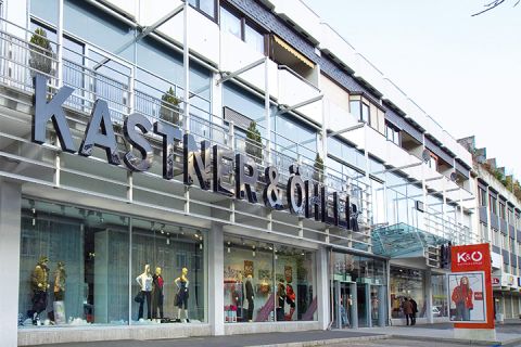 Das Bild zeigt die Ladenfront von Kastner & Öhler mit einem großen rot-weißen Logo auf der linken Seite und einer modernen Glasfassade. In den Fenstern unter dem markanten Namensschild des Ladens stehen Schaufensterpuppen in stilvoller Kleidung.