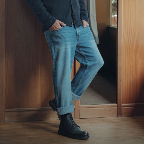 Eine Person steht in einem Haus mit den Händen in den Taschen, trägt eine blaue Jeans mit Manschetten, eine dunkle Jacke und schwarze Schnürstiefel. Nur die untere Hälfte ihres Körpers ist sichtbar, sie lehnt an einer Holzwand.