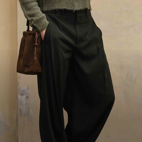 Eine Person, die eine schwarze Hose mit weitem Bein, einen olivgrünen Pullover und eine braune Wildleder-Kordeltasche trägt, steht mit einer Hand in der Tasche an einer beigen Wand.