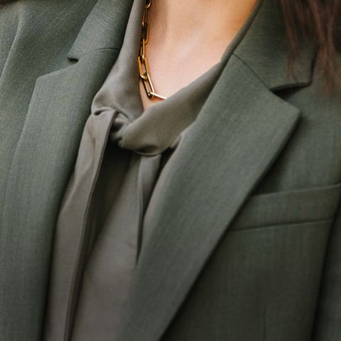 Nahaufnahme einer Person, die einen olivgrünen Blazer und eine dazu passende Bluse mit einer Schleife am Kragen trägt, die mit einer klobigen Goldkette verziert ist.