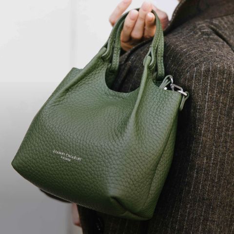 Eine Person, die einen braunen Nadelstreifenmantel trägt, hält eine strukturierte grüne Lederhandtasche mit kurzen Griffen. Auf der Vorderseite der Tasche ist in weißen Buchstaben "GIANNI CHIARINI FIRENZE" aufgedruckt.