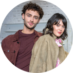 Ein Mann und eine Frau stehen dicht beieinander, beide tragen legere Jacken, vor einer weißen Backsteinmauer und schauen in die Kamera.