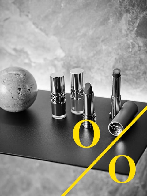 Vier Lippenstifte in Metalltuben auf einer dunklen Fläche, mit zwei gelben Buchstaben "o" und einer diagonalen gelben Linie im Vordergrund. Schwarz-weißes Bild.