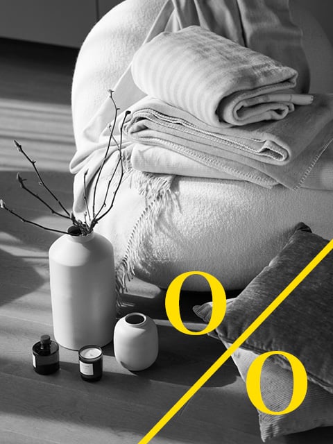 Eine gemütliche Szene mit gefalteten Decken, einem Sitzsack, Vasen mit Zweigen und Kerzen auf einem Holzboden bei natürlichem Licht.