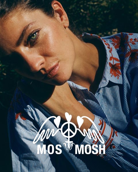 Eine Frau mit langem, gewelltem blondem Haar trägt ein grünes Hemd mit weißen Mustern. Sie blickt über ihre Schulter und steht vor einem modernen Gebäude. Das "MOS MOSH"-Logo befindet sich in der oberen linken Ecke.