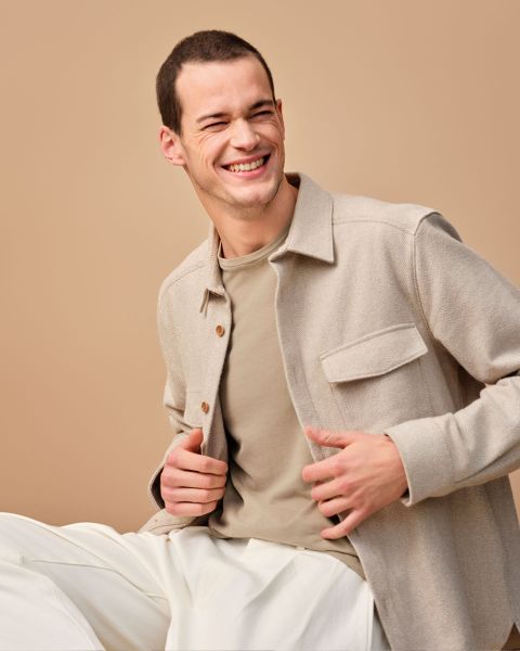 Lächelnder Mann sitzt vor einem beigen Hintergrund, trägt eine hellbraune Jacke über einem hellbraunen Hemd und weißen Hosen und blickt mit fröhlichem Gesichtsausdruck zur Seite.