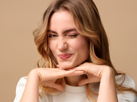 Eine Person mit langem, welligem Haar und leichtem Make-up lächelt mit geschlossenem Auge und stützt ihr Kinn auf die Hände. Sie trägt ein weißes Oberteil vor einem einfarbig beigen Hintergrund.