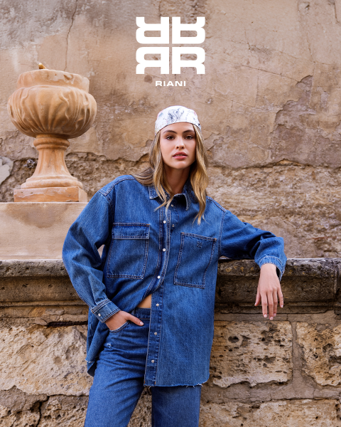 Zwei Frauen posieren stilvoll in einem Theater mit roten Sitzen. Die eine sitzt auf der Rückenlehne eines Sitzes und trägt ein gemustertes Kleid, einen schwarzen Mantel und Stiefel. Die andere steht in einem marineblauen Pullover und einem schwarzen Rock neben ihr. Über ihnen erscheint das Logo "RIANI".
