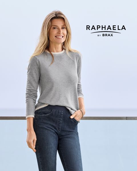Eine Frau mit langen blonden Haaren, die ein graues langärmeliges Hemd und dunkle Jeans trägt, steht lächelnd an einem Geländer mit hellblauem Hintergrund. Neben ihr erscheint der Text "EUREX by BRAX".