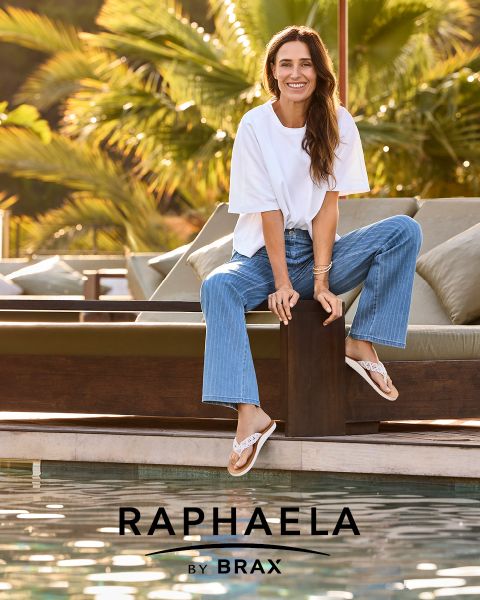 Eine Frau mit langen blonden Haaren, die ein graues langärmeliges Hemd und dunkle Jeans trägt, steht lächelnd an einem Geländer mit hellblauem Hintergrund. Neben ihr erscheint der Text "EUREX by BRAX".