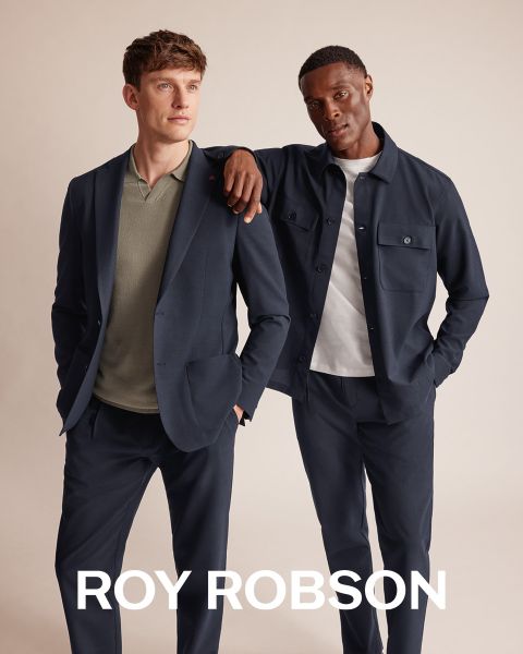 Ein Mann steht im Freien vor verschneiten Bergen und trägt einen grauen Hahnentrittmantel, einen grauen Pullover und eine graue Hose. Der Text "ROY ROBSON" erscheint am unteren Rand des Bildes.