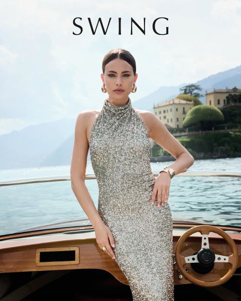 Eine Frau in einem seidigen, salbeigrünen Kleid sitzt auf einem cremefarbenen Stuhl vor einem floralen Hintergrund. Sie trägt goldene Absätze, Statement-Ohrringe und ein Armband. Das Wort "SWING" erscheint am unteren Rand des Bildes.