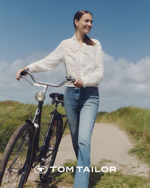 Eine lächelnde Frau in einem kastanienbraunen Pullover und ein Mann in einem marineblauen Pullover stehen Seite an Seite an einem Strand. Beide tragen Jeans. Der Himmel ist klar und blau. Das Logo "Tom Tailor" erscheint am unteren Rand.