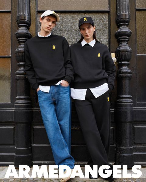 Zwei Personen tragen passende schwarze Sweatshirts mit einem gelben Bandemblem und legere Hosen, eine in Jeans und die andere in Schwarz. Sie stehen vor einer dunklen Wand. Unten ist das Wort „ARMEDANGELS“ zu sehen.