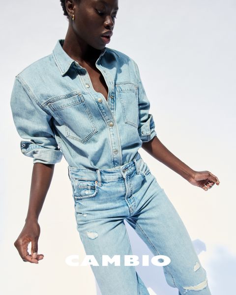 Person, die ein helles Hemd und Jeans trägt und vor einem schlichten Hintergrund posiert. Unten ist das Wort „CAMBIO“ zu sehen. Das Outfit wirkt lässig und stilvoll.