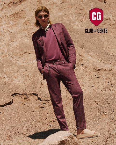 Mann in burgunderfarbenem Anzug und Sonnenbrille steht auf felsigem Gelände, lehnt sich zurück und hat die Hände in den Taschen. Logo: „Club of Gents“ in der oberen rechten Ecke.