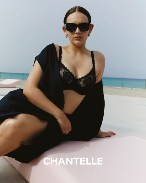 Eine Frau mit Sonnenbrille und schwarzem Spitzen-BH sitzt auf einer rosa Fläche am Strand. Sie ist in ein schwarzes Kleidungsstück gehüllt, im Hintergrund ist das Meer zu sehen. Unten ist das Wort „CHANTELLE“ zu sehen.