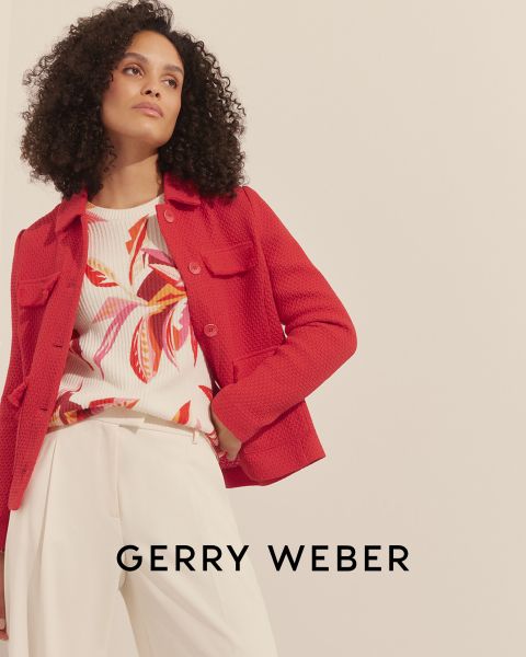 Eine Frau mit lockigem Haar trägt eine rote Strukturjacke über einem bunten geblümten Oberteil und cremefarbenen Hosen. Der Hintergrund ist schlicht und unten ist der Text „Gerry Weber“ zu sehen.