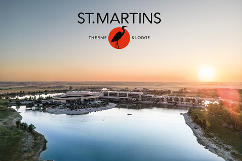 Luftaufnahme der St. Martins Therme & Lodge an einem See bei Sonnenuntergang, mit dem Logo und einer Vogelsilhouette über den Gebäuden.